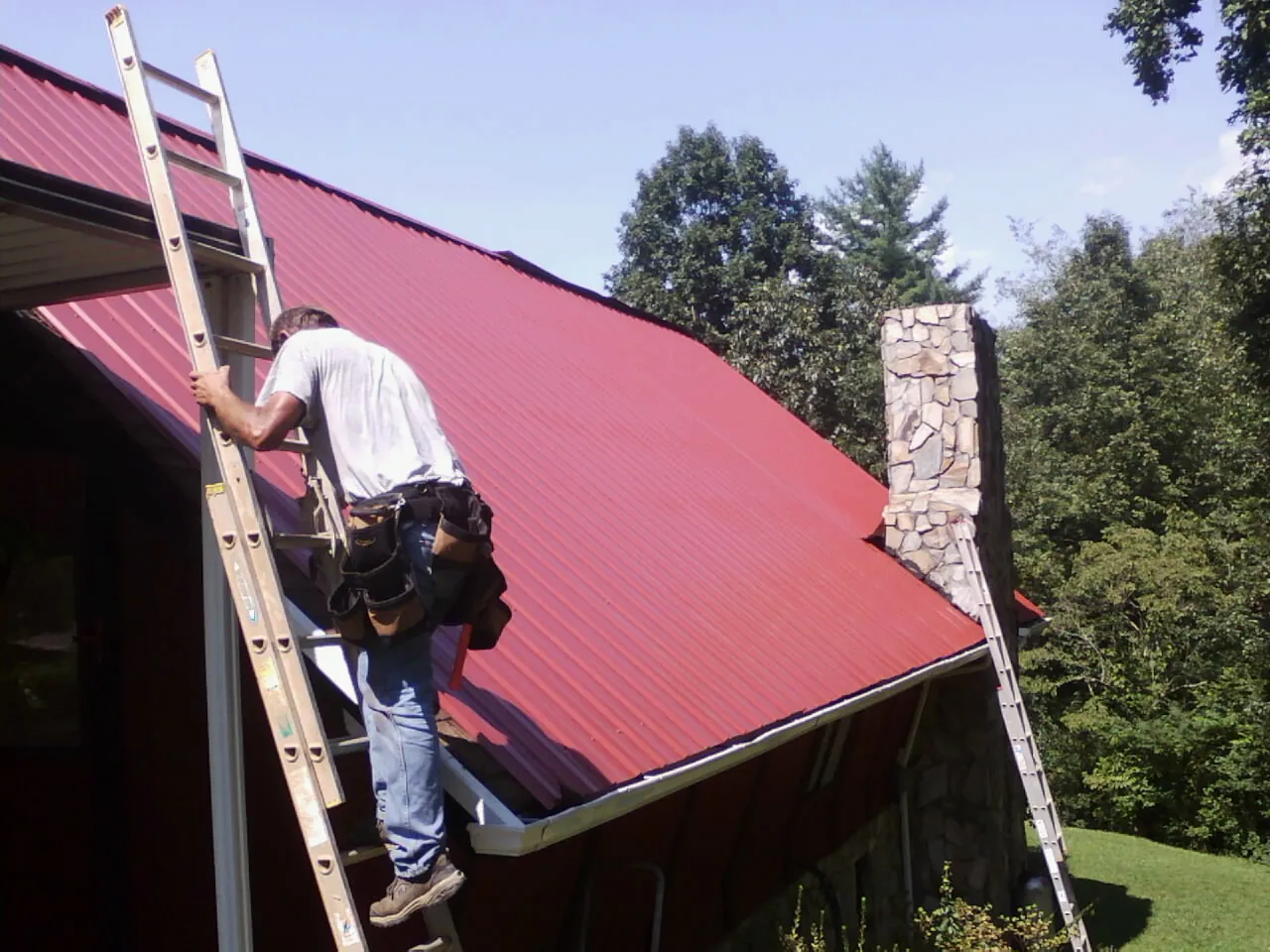 Roofer on a ladder beside a red metal roof and stone chimney — Soffit & Fascia Repair by Roofing Brothers in Decatur